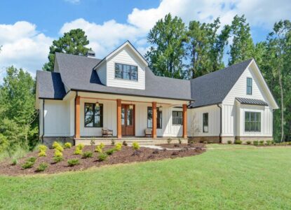 A charming white farmhouse featuring a spacious front porch, surrounded by greenery and a serene landscape.